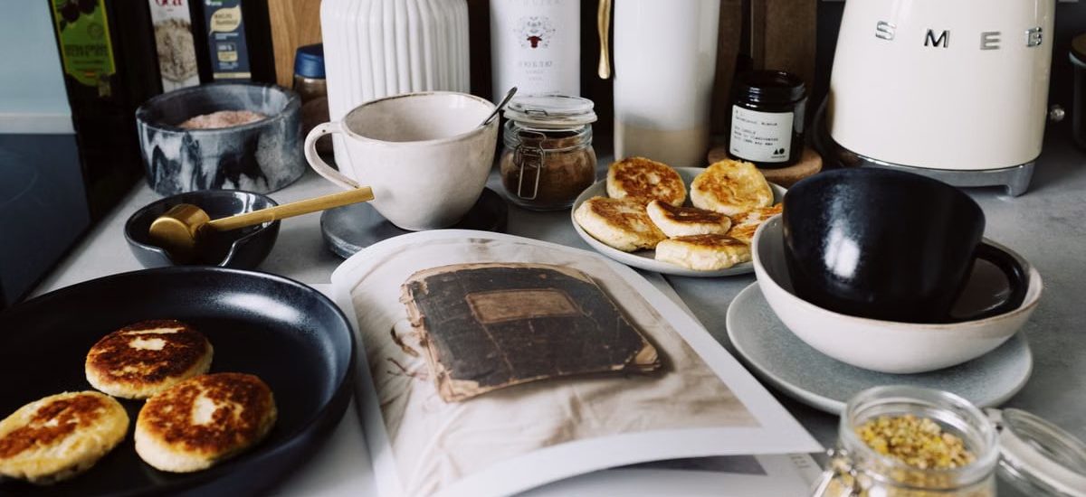 One portion of homemade cheese pancakes in frying pan and another on plate locating near opened stylish magazine among kitchen utensils and appliances in bright kitchen at home