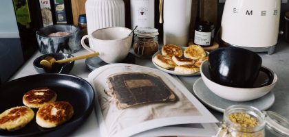 One portion of homemade cheese pancakes in frying pan and another on plate locating near opened stylish magazine among kitchen utensils and appliances in bright kitchen at home