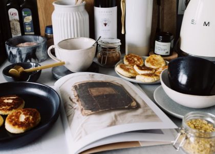 One portion of homemade cheese pancakes in frying pan and another on plate locating near opened stylish magazine among kitchen utensils and appliances in bright kitchen at home