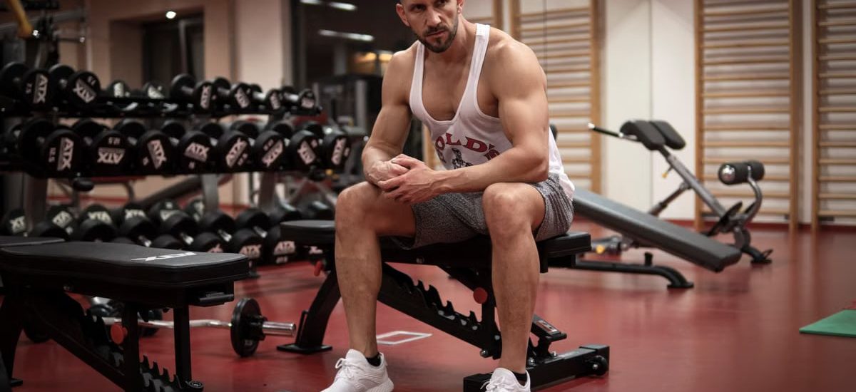 Strong, determined man in white tank top sitting in gym, showcasing fitness and strength.