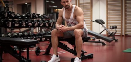 Strong, determined man in white tank top sitting in gym, showcasing fitness and strength.